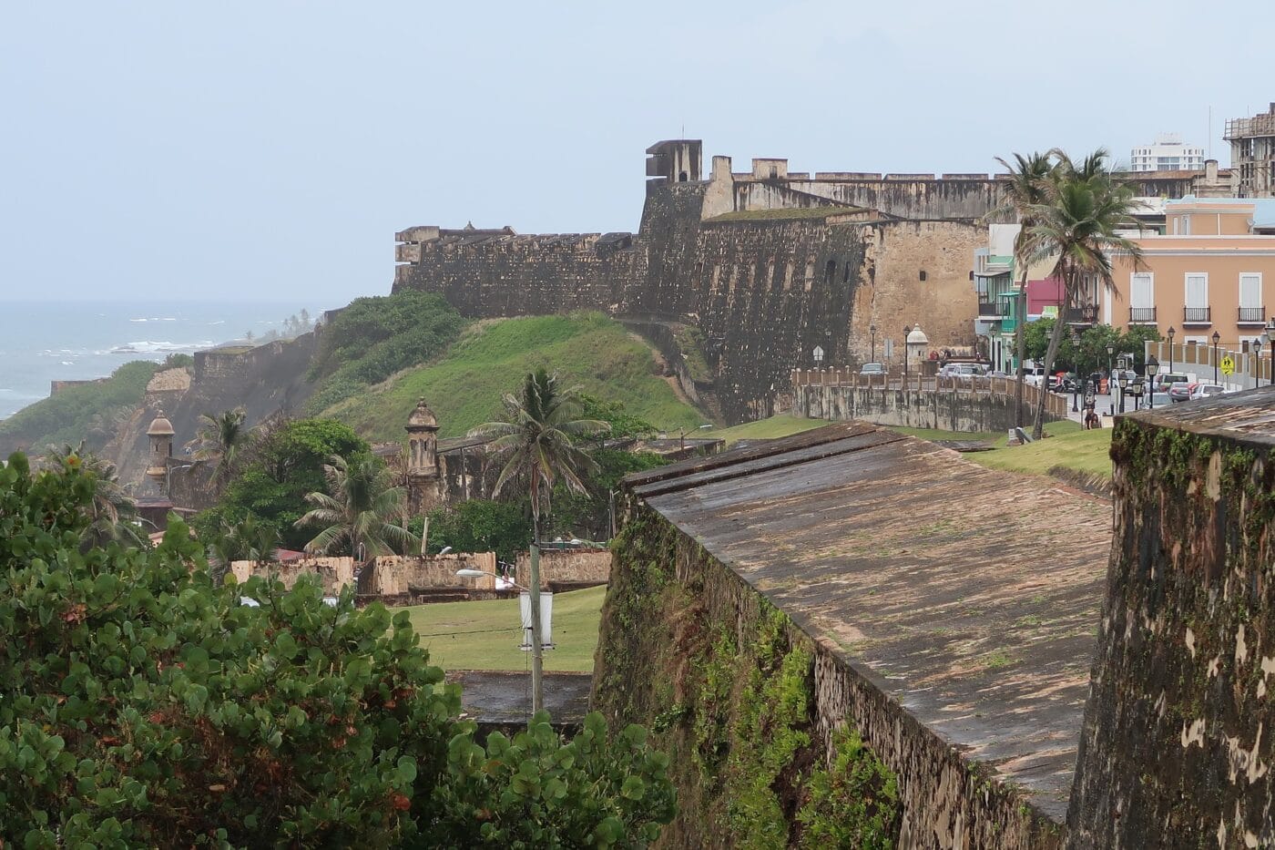 Ground-level view of the historic El Morro fortress in San Juan, Puerto Rico, with its massive stone walls and lush greenery surrounding the structure. Palm trees and colorful buildings are visible nearby, along with the ocean in the background under a cloudy sky.