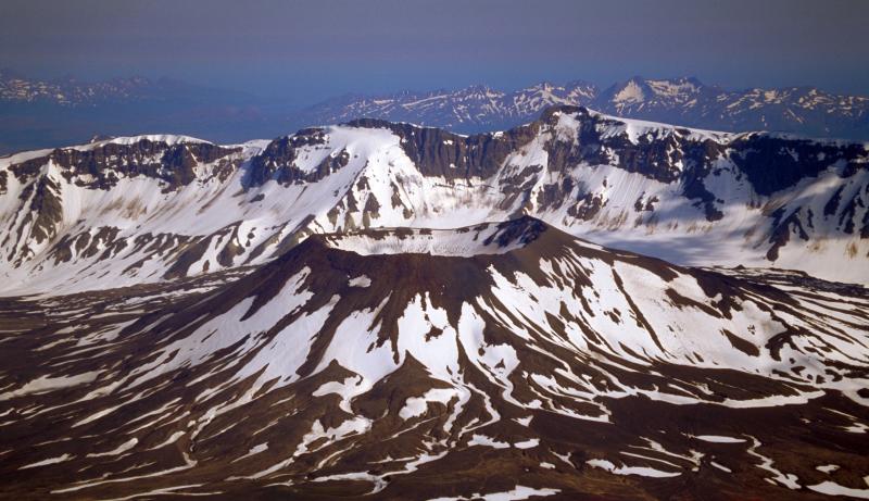 A snow covered small volcano and a mountain range behind it. Aniakchak National Monument