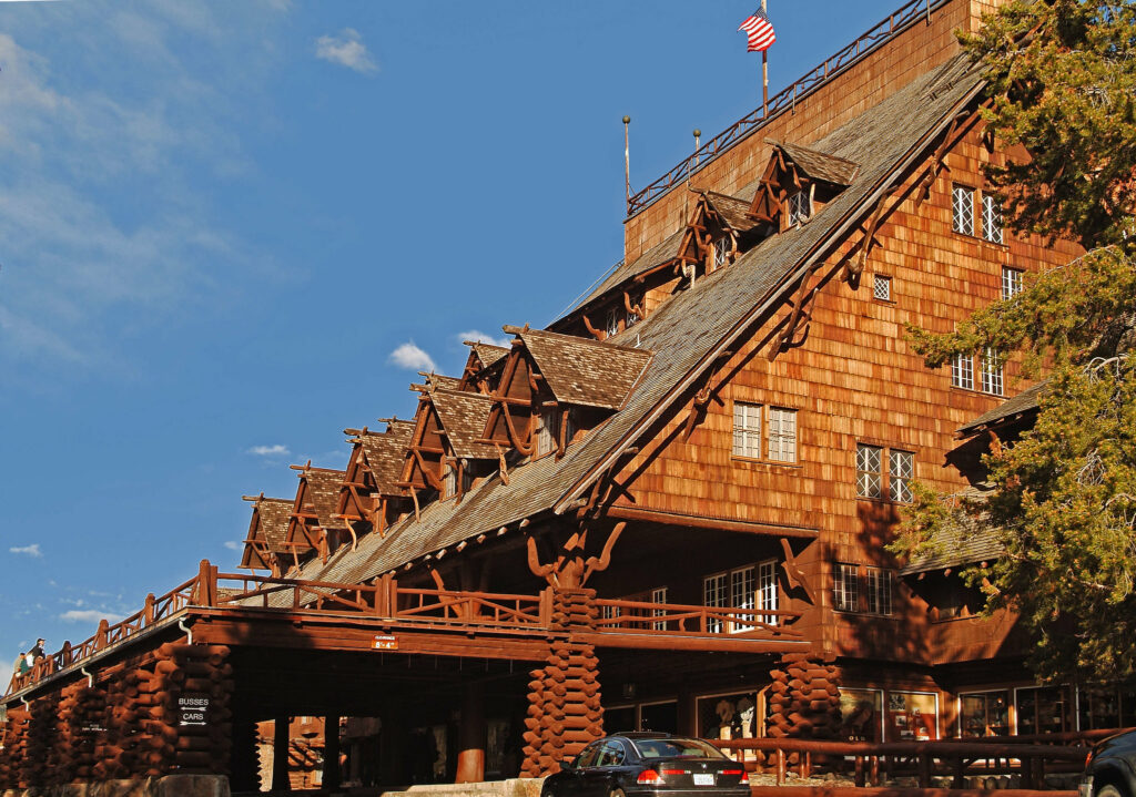 A massive historic lodge with reddish brown wooden shingles, steeply pitched roofs, and multiple dormer windows. An American flag flies above the rustic building against a bright blue sky.