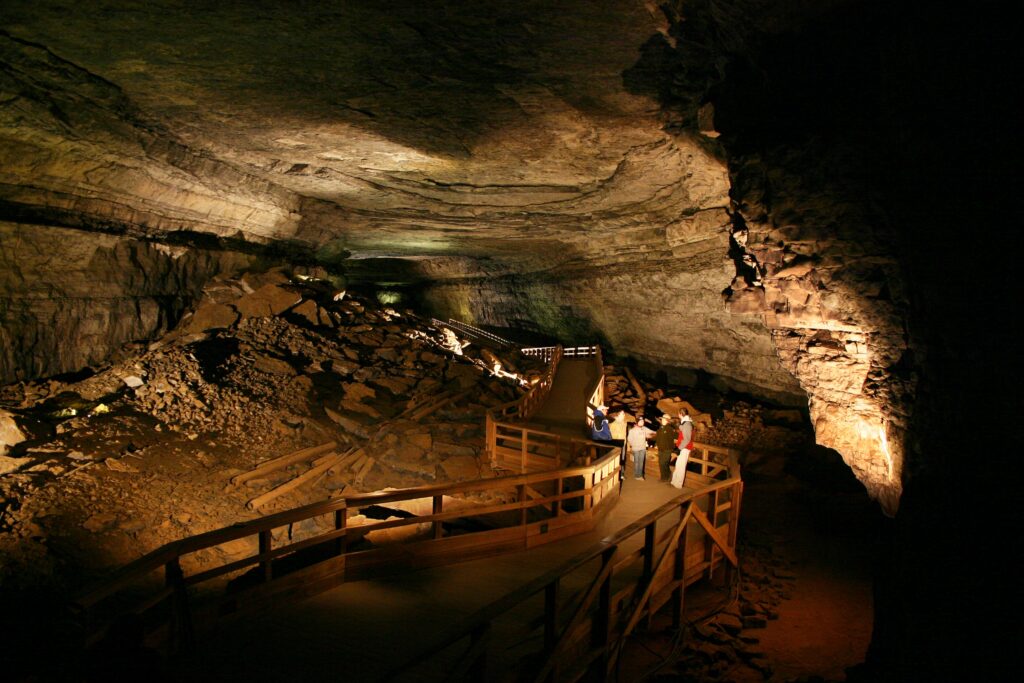 A dimly lit underground cavern with a wooden boardwalk winding between massive rock formations. Several visitors stand along the walkway beneath the low stone ceiling.