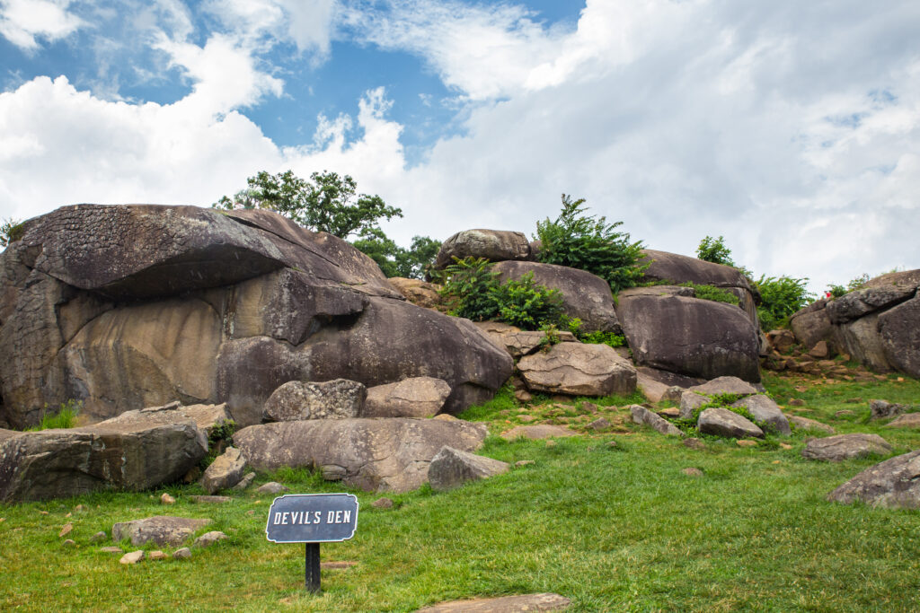 Massive rounded granite boulders form rocky crevices at Devils Den in Gettysburg National Military Park. A small sign reading "DEVIL'S DEN" stands in the grass in front of the historic battlefield landmark.