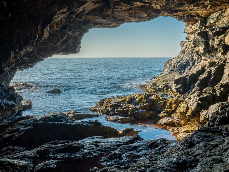 View from inside a dark rocky sea cave looking out toward the blue ocean through a naturally formed opening. Sunlight illuminates the jagged rocks and tidal pools along the cave floor, creating an atmospheric scene for a haunted national park story.