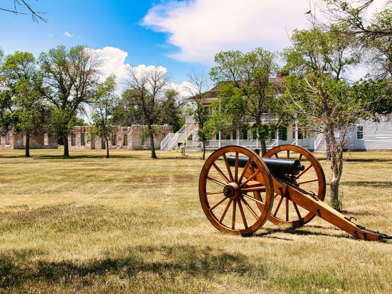 A historic black cannon mounted on a wooden carriage stands on a grassy lawn in front of white buildings and stone ruins. Mature trees surround the preserved military site beneath a bright blue sky.