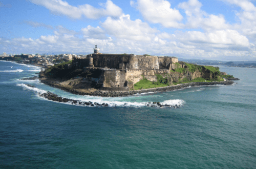Aerial view of El Morro, a historic fortress located in San Juan, Puerto Rico, surrounded by the Atlantic Ocean with waves crashing against the rocky coastline, under a partly cloudy sky.