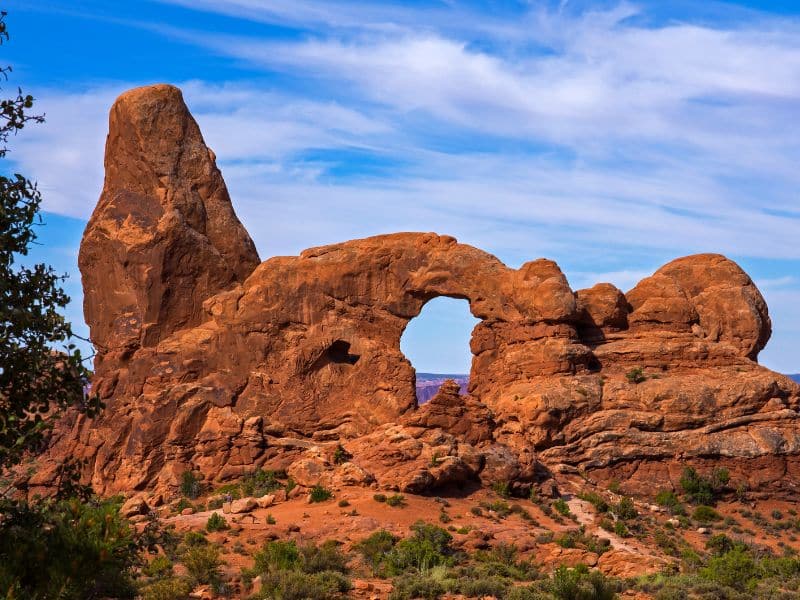 Red sandstone rock formations under a blue sky in Arches National Park, showcasing a prominent natural arch and spire, highlighting the park's unique geological features. The image captures the stunning landscape and iconic rock formations visitors can expect when visiting Arches National Park.