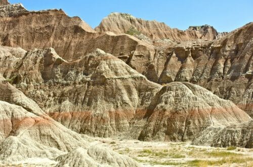 Eroded rock formations with distinct layers of sediment under a clear blue sky in Badlands National Park, illustrating the park's unique and rugged landscape. The image highlights the dramatic scenery and geological features visitors can explore when visiting Badlands.