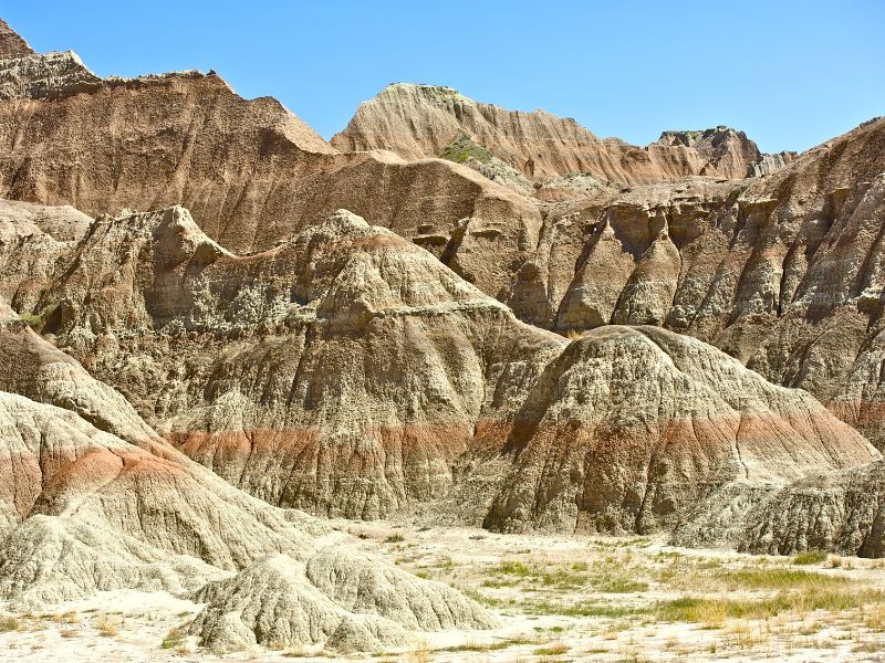 Eroded rock formations with distinct layers of sediment under a clear blue sky in Badlands National Park, illustrating the park's unique and rugged landscape. The image highlights the dramatic scenery and geological features visitors can explore when visiting Badlands.