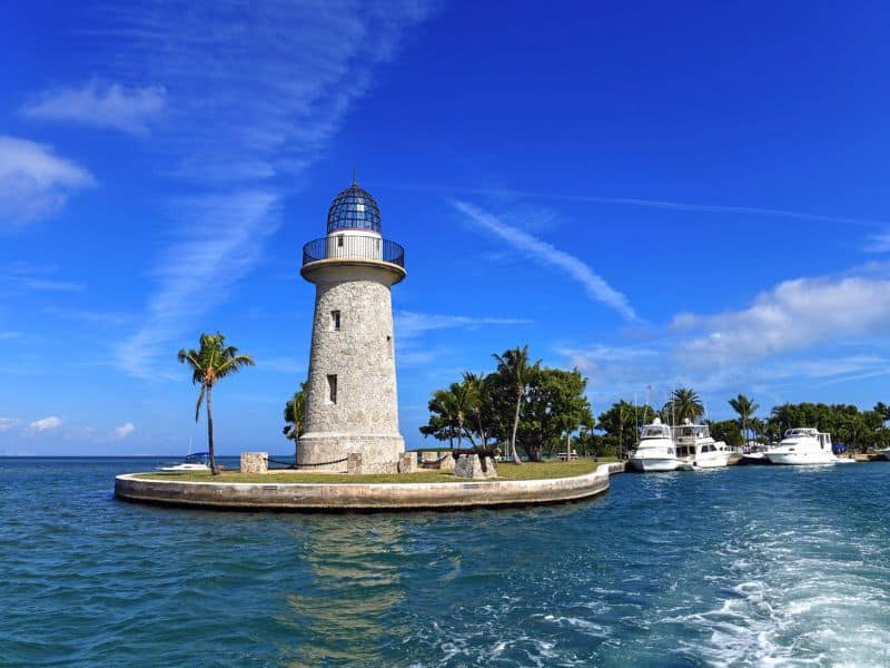 The image depicts a lighthouse on a small, circular island with a few palm trees, surrounded by blue water under a clear, vibrant sky. Nearby, several white yachts are docked along the shoreline, indicating a marina. The scene is peaceful and picturesque, with the lighthouse standing prominently as a focal point.