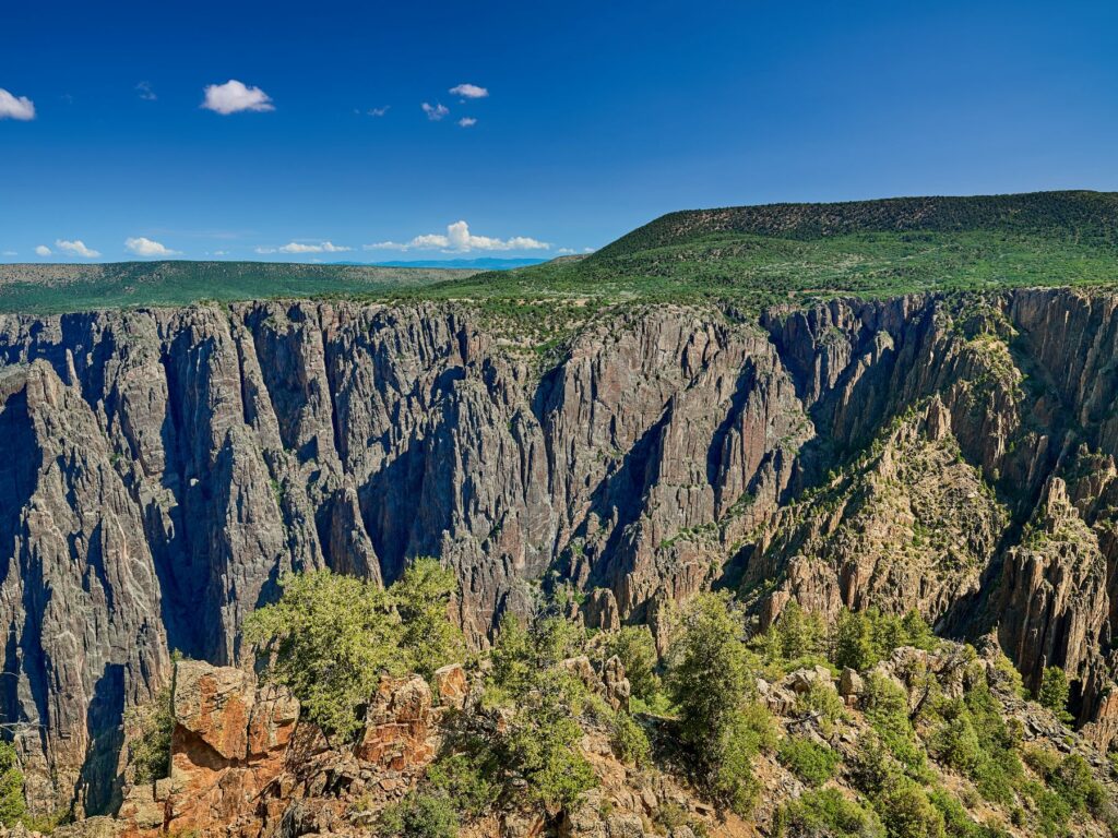 The image shows the dramatic cliffs of Black Canyon of the Gunnison, with sheer rock faces plunging into the deep, narrow canyon below. The rugged terrain is highlighted by patches of green vegetation, and the expansive view under a clear blue sky showcases the vastness and rugged beauty of the landscape.