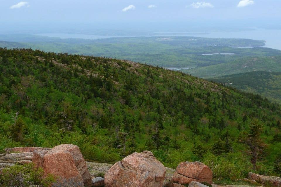 Scenic view from the summit of a hill in Acadia National Park, overlooking a lush landscape of dense, green forests stretching toward a distant coastline. The foreground features rugged, rounded boulders typical of the area. In the distance, the waters of the Atlantic Ocean merge with the overcast sky, creating a serene and misty atmosphere. This image captures the natural beauty and diverse terrain of the park.