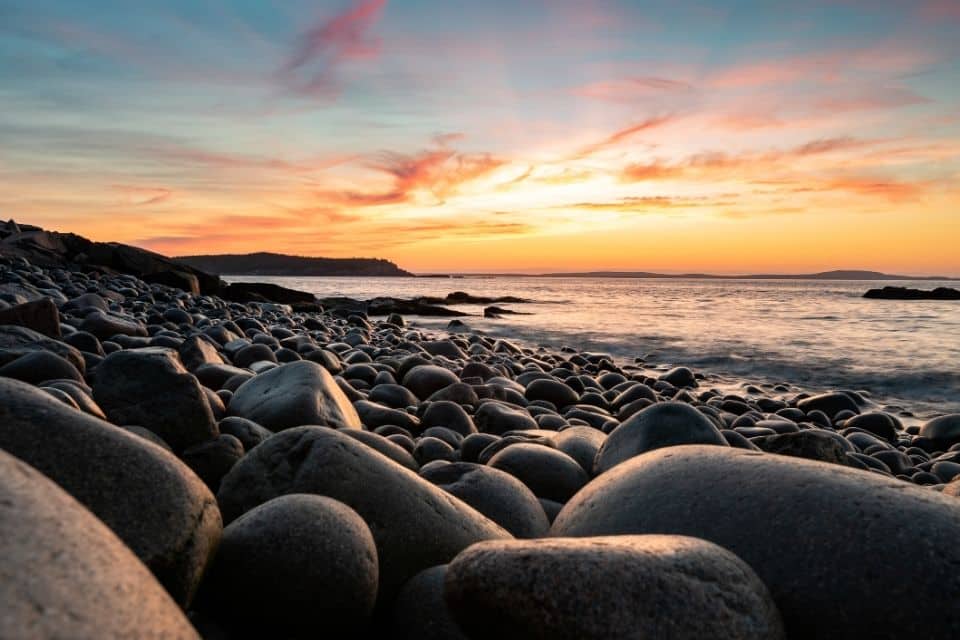 Breathtaking sunset at Acadia National Park with a foreground of smooth, rounded beach stones leading to gentle ocean waves. The sky displays vibrant shades of orange, pink, and blue above the serene coastline. The peaceful scene captures the natural beauty and tranquil atmosphere of the park at dusk.