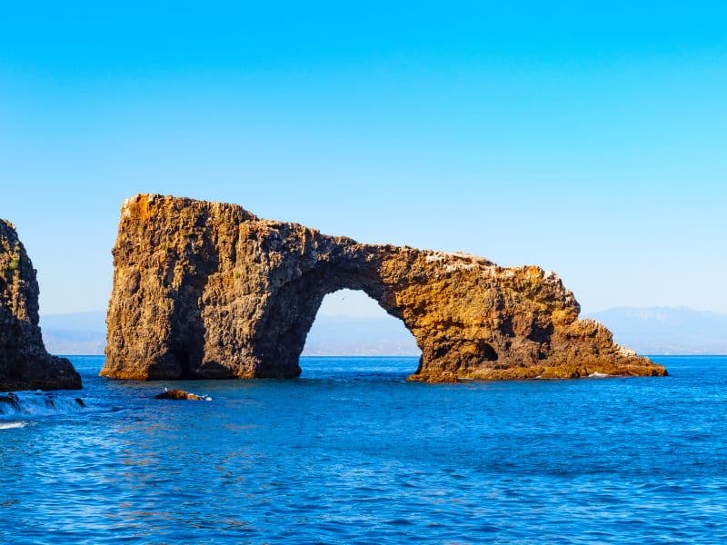 The image shows a natural rock arch formation standing in the clear blue waters of the ocean, with a backdrop of distant mountains under a bright blue sky. The arch, made of rugged, weathered rock, creates a striking and iconic coastal scene. The calm water enhances the serene beauty of the landscape.
