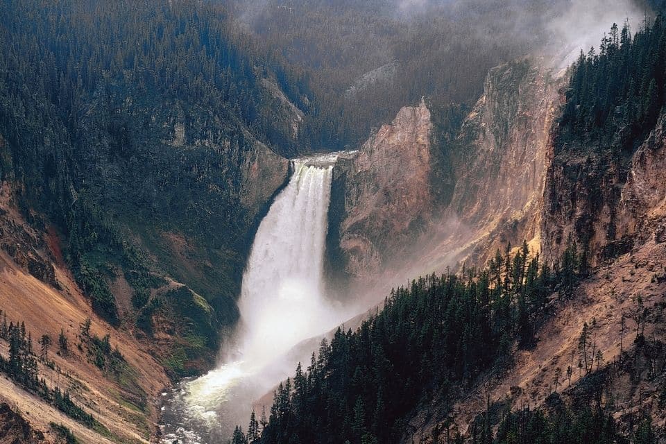A dramatic view of the Lower Falls of the Yellowstone River, where a powerful waterfall cuts through a rugged canyon shrouded in mist, surrounded by steep cliffs and dense forests.