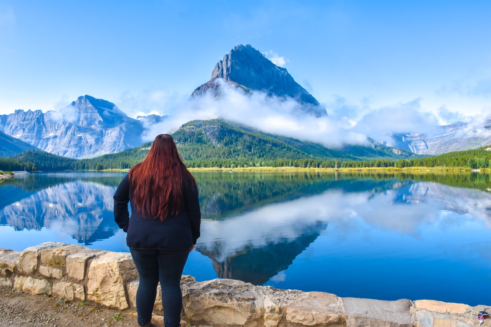 Woman standing next to a mountain partially hidden by clouds reflected by a lake in Many Glacier, Glacier National Park