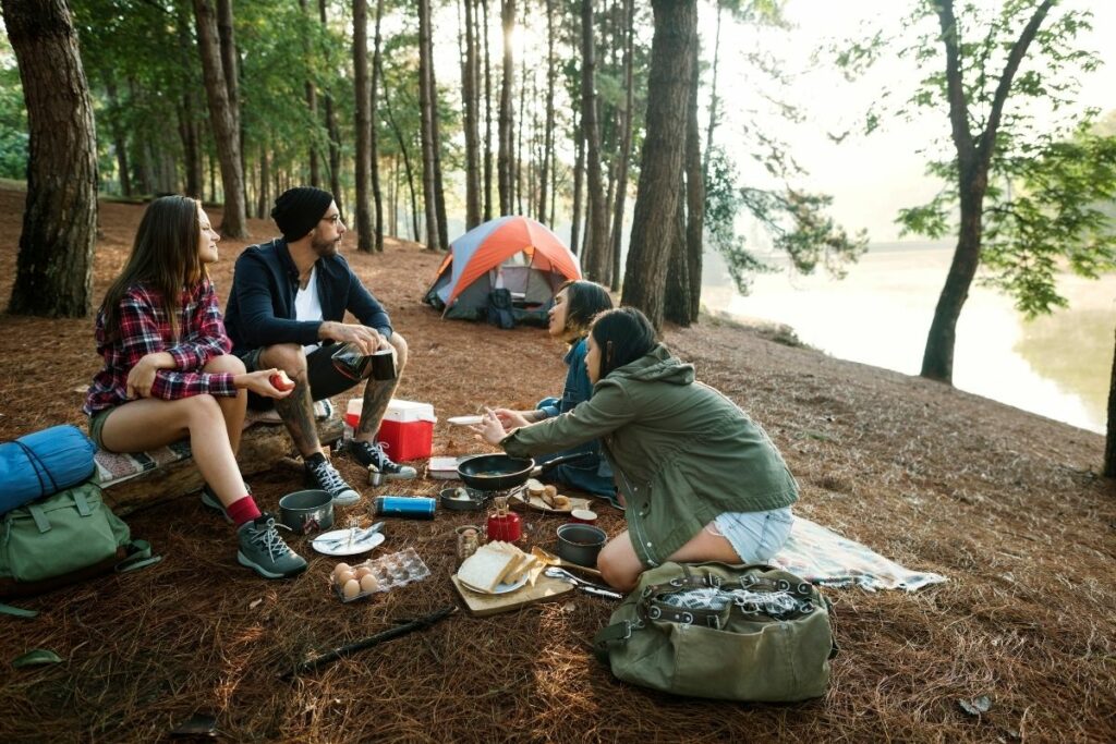 3 women and a man having breakfast while camping by a lake. 
