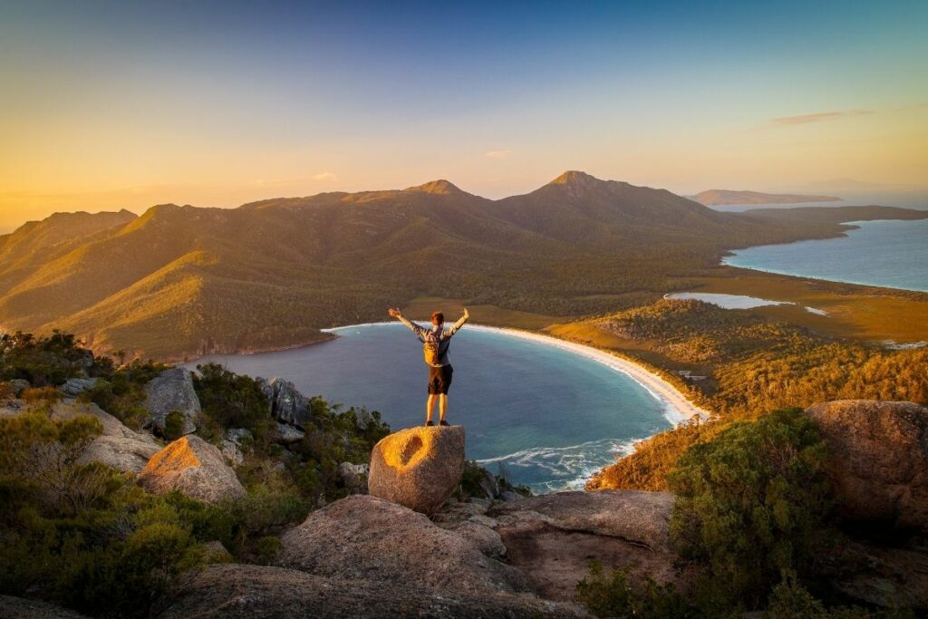 A man standing with his arms raised enjoying the view of the mountains and a bay.  
