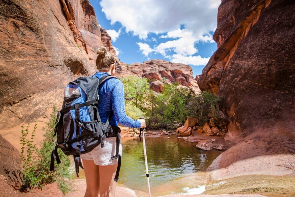 A women in a blue shirt with trekking poles near a pool of water in a Utah Canyon