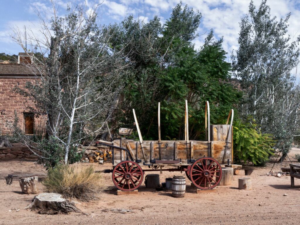 A weathered wooden wagon with red-spoked wheels stands in a rustic desert yard, surrounded by trees and an old brick building in the background.
