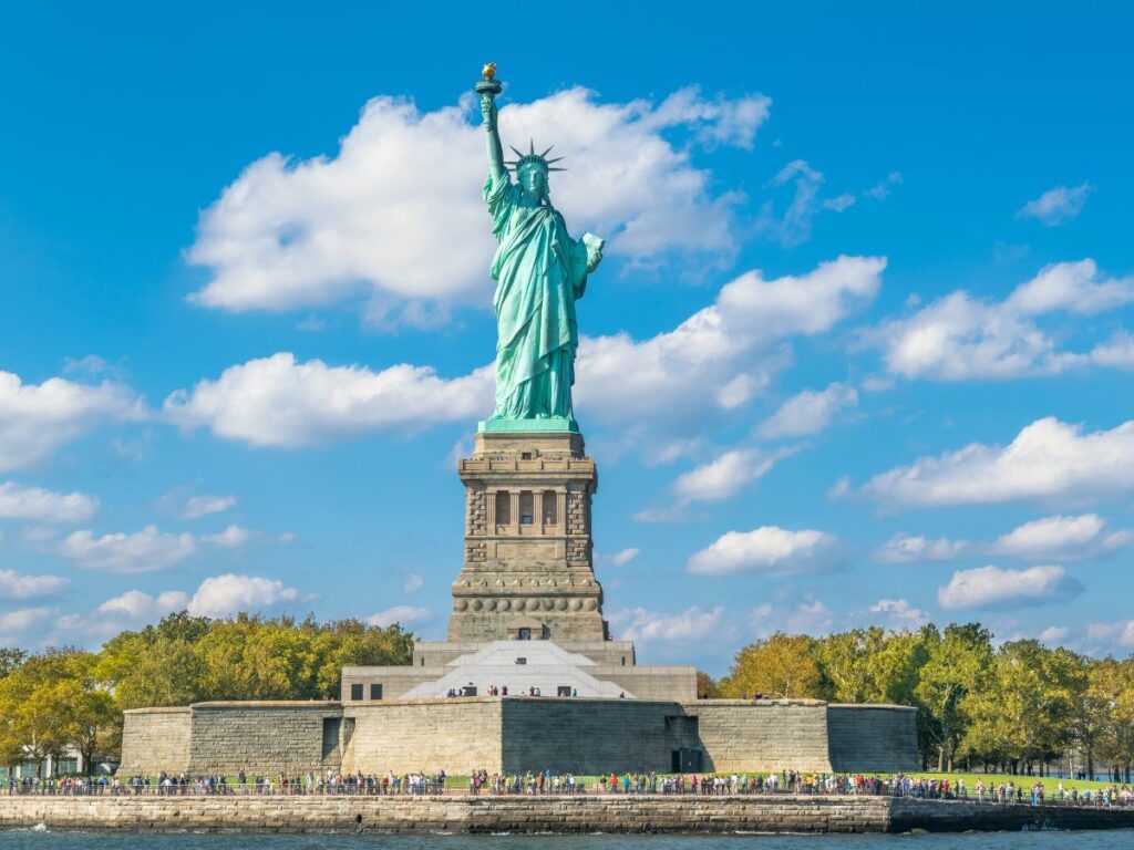 The Statue of Liberty stands tall against a bright blue sky with scattered clouds, overlooking a crowd of visitors at its base on Liberty Island. The iconic green copper statue holds a torch high in her right hand and a tablet in her left.