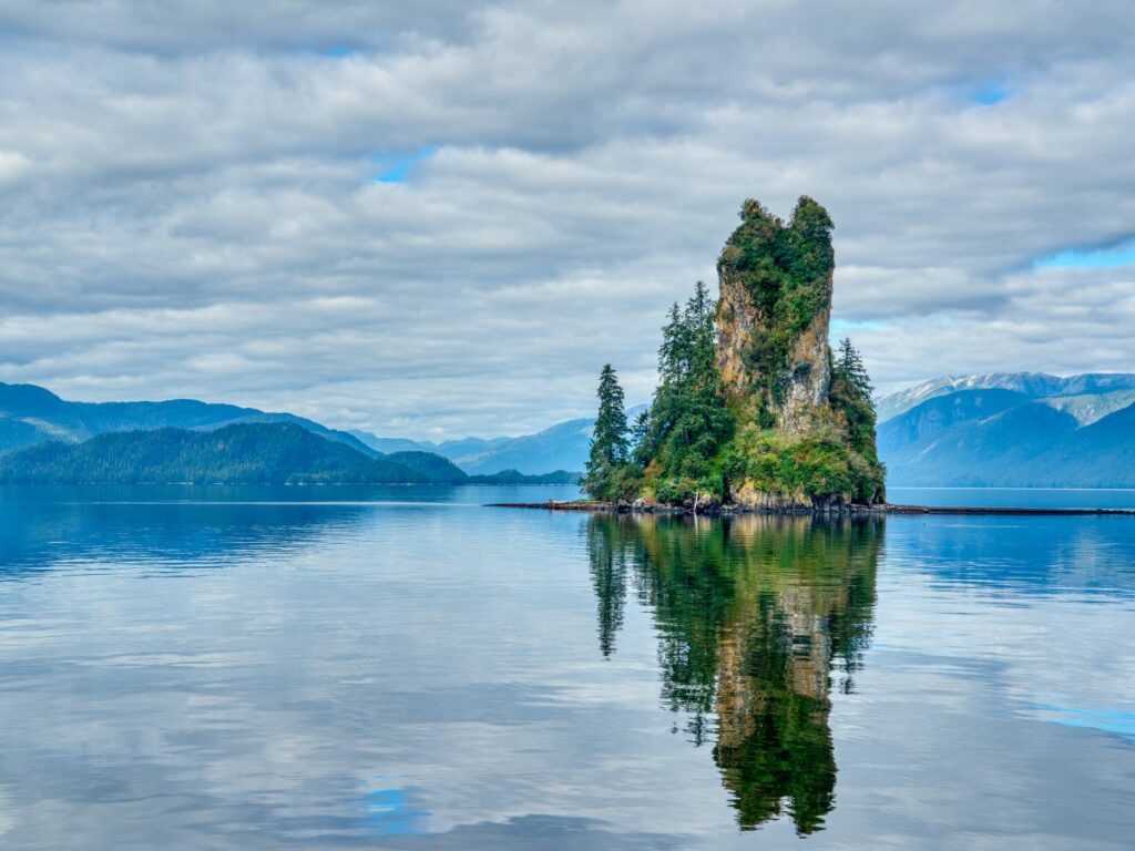 A small, lushly forested sea stack rises from calm blue water, perfectly reflected on the surface, with forested mountains and a cloudy sky in the background.