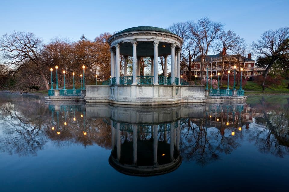 Twilight scene of a neoclassical gazebo with teal trim, reflected in the still water of a pond, with lit lamps casting a warm glow. In the background, a historic mansion with illuminated windows is nestled among bare autumn trees.