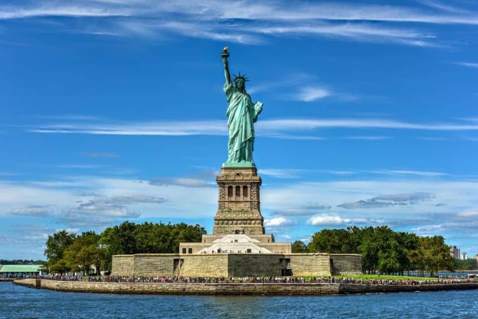The Statue of Liberty stands tall against a clear blue sky with scattered clouds. This iconic symbol of freedom is centered in the image, with its green patina contrasting with the bright sky. The statue is on its traditional stone pedestal, and below, crowds of visitors can be seen walking around the island's perimeter. The water surrounding the island is calm, reflecting the light of a sunny day. Trees flank the left side, adding a touch of greenery to the scene.