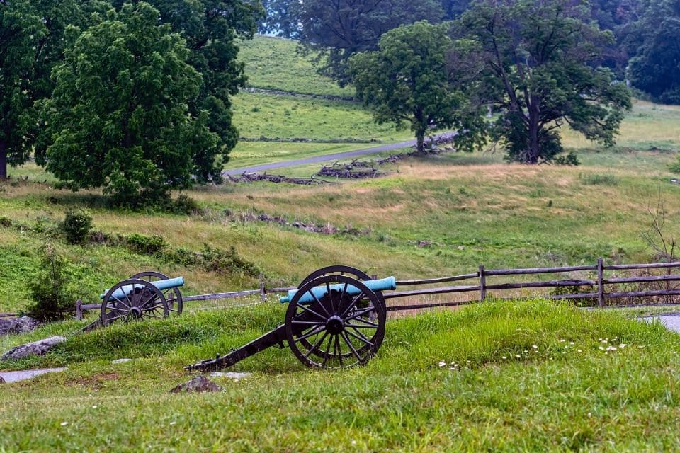 A historic battlefield with two cannon displayed in the foreground, overlooking a vast, gently rolling landscape with split-rail fences and mature trees. The overcast sky and the dampness of the green grass suggest a quiet, reflective atmosphere on this former ground of conflict.