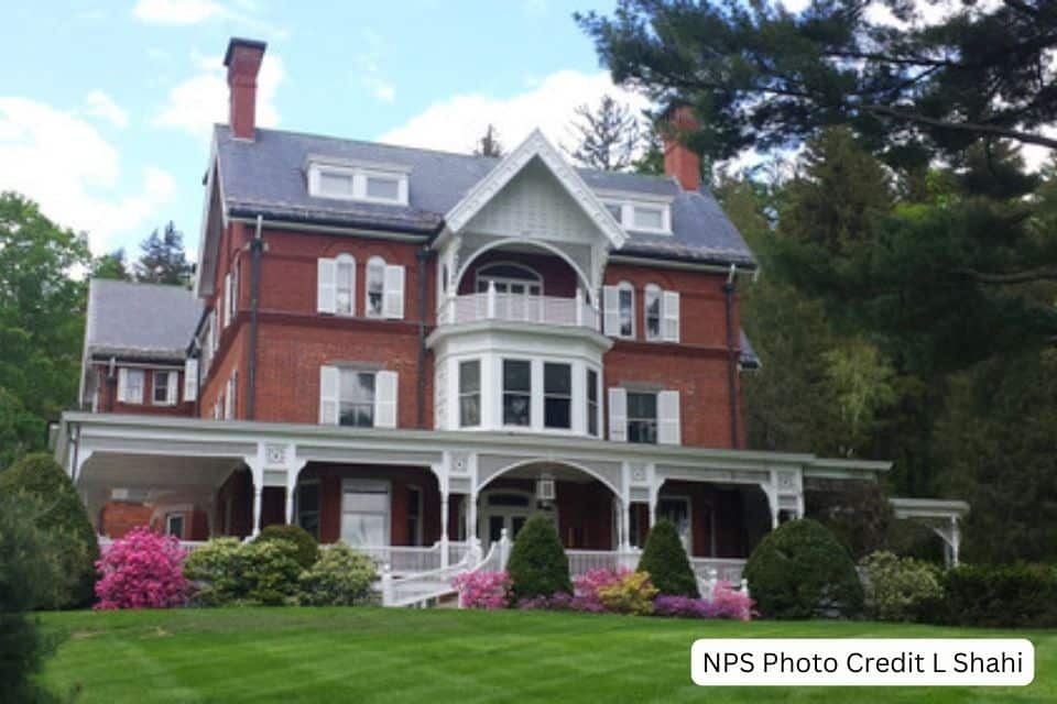 A majestic Queen Anne style red brick mansion with a rounded front turret, white trim, and a wide porch surrounded by lush green lawns and blooming pink rhododendron bushes. The home's historic character is accented by tall chimneys and a backdrop of mature trees under a partly cloudy sky.