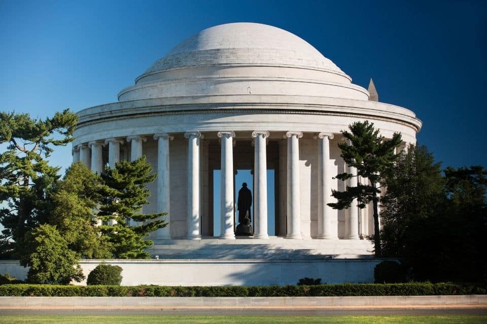 The Thomas Jefferson Memorial in Washington D.C., a neoclassical structure with a shallow dome, surrounded by a circular colonnade of Ionic columns. A statue of Jefferson stands in the center, visible through the open doorway, with greenery framing the monument under a clear blue sky.