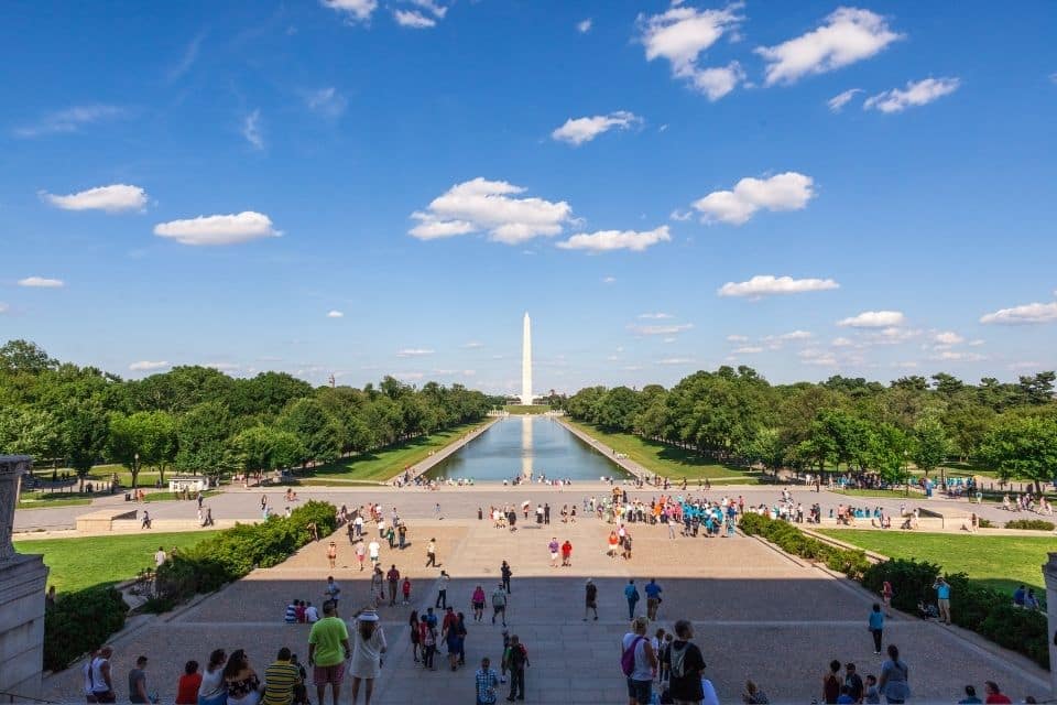 A vibrant scene from the Lincoln Memorial, looking out over the Reflecting Pool towards the Washington Monument in the distance. The sky is blue with scattered clouds, and the area is bustling with tourists and visitors enjoying the panoramic view and the serene atmosphere of the National Mall.