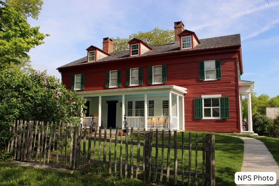 A traditional two-story colonial house with red walls and white trim, a covered front porch, and a black shingled roof. A wooden picket fence surrounds the front yard, which is lush with green grass and flowering shrubs under a clear blue sky.