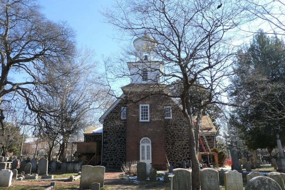 An old stone church with a unique rounded stone facade and a white wooden steeple, situated in a graveyard with tombstones dating back centuries. Bare trees frame the building under a clear winter sky.