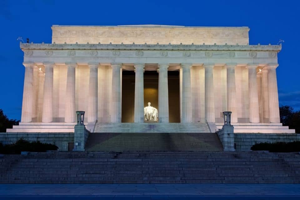 The Lincoln Memorial at dusk, illuminated from within, showcasing the grandeur of its neoclassical architecture with strong columns and a sculpted frieze. The statue of Abraham Lincoln is visible in the central hall, seated and contemplative against the softly lit marble interior.