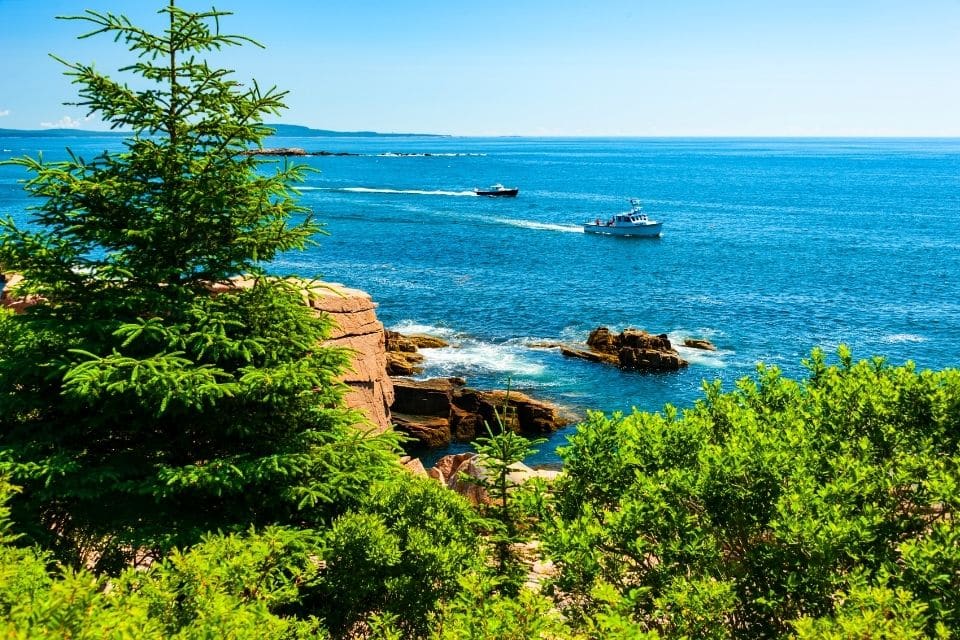 A picturesque coastal scene with rugged, rocky shores and lush greenery in the foreground. A clear blue sky hangs over calm ocean waters, where two boats are sailing near the coastline, creating a serene maritime landscape.
