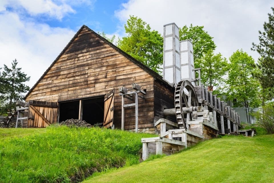 A historic wooden barn with a large water wheel attached to its side, set in a green field. The barn's rustic appearance and the functional water wheel suggest a bygone era of agriculture, against a backdrop of lush trees and overcast skies.