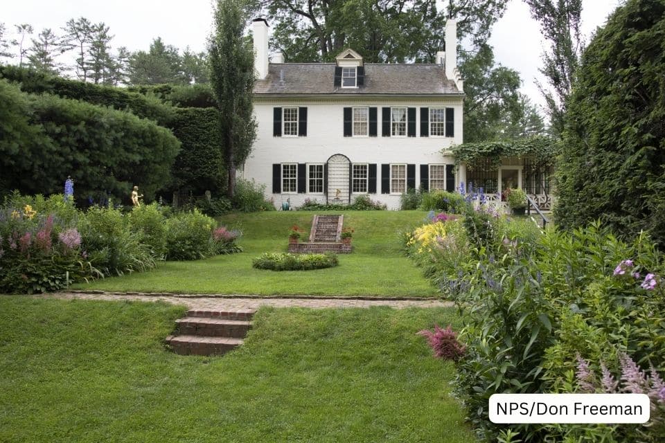 A classic white colonial house with green shutters, surrounded by a well-manicured garden full of colorful flowers and shrubs. A brick pathway leads to the front steps, creating a welcoming atmosphere in this idyllic, tranquil setting.