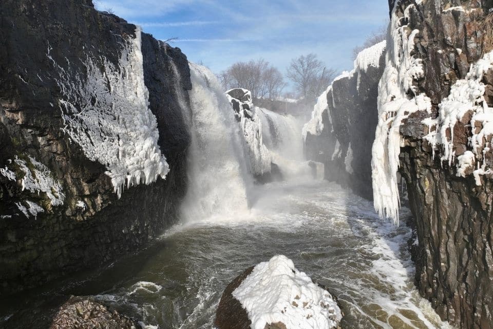 A dramatic waterfall partially frozen over with large icicles and snow, surrounded by steep dark cliffs. The powerful cascade of water cuts through the ice, creating a dynamic and contrasting winter scene.