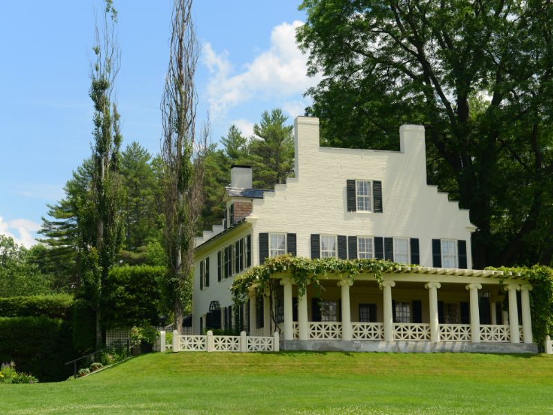 A white historic house with black shutters and a vine covered porch sits on a manicured green lawn surrounded by tall trees.