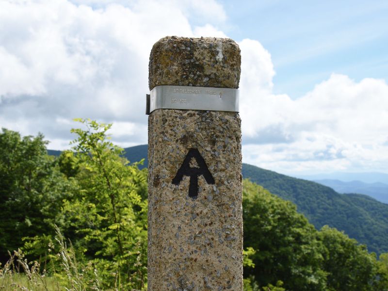 An Appalachian Trail marker with the black AT symbol stands at a scenic mountain overlook beneath a blue and cloudy sky. The forested mountain landscape highlights the hiking opportunities found in New Hampshire.