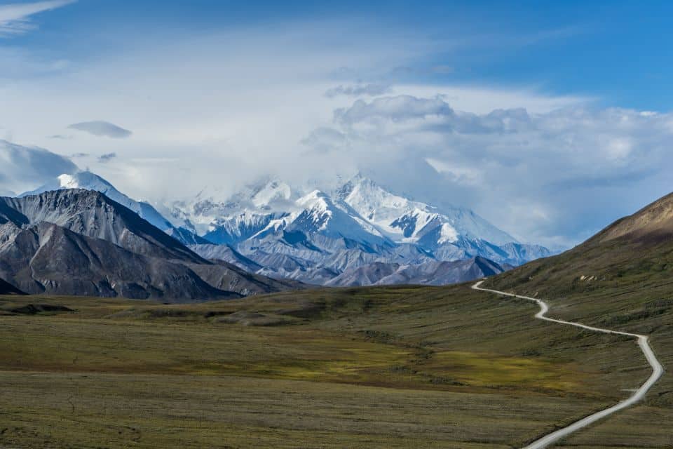 A winding road meanders through a vast, grassy tundra towards majestic, snow-capped peaks shrouded in clouds, showcasing the serene and rugged landscape of Denali National Park.