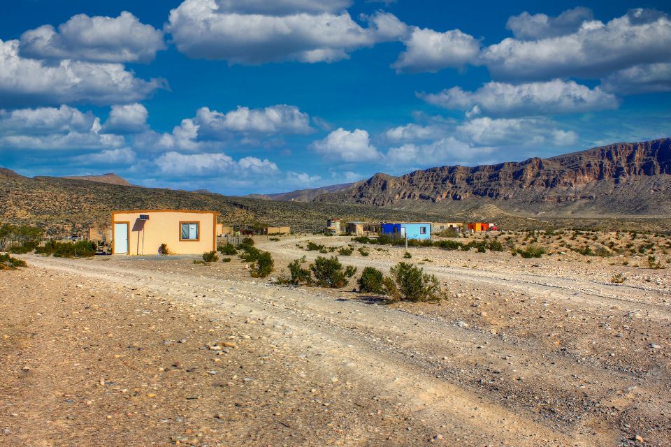 Colorful, isolated desert homes with flat roofs stand in a sparse, rocky landscape with mountains in the distance and a bright blue sky dotted with fluffy white clouds.