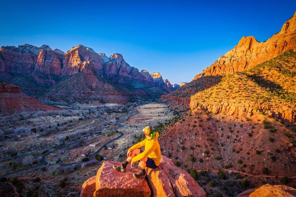 An adventurer rests on a red sandstone cliff at sunset in Zion National Park, showcasing a breathtaking view ideal for 'Hiking Tips for Beginners' on timing and scenery.
