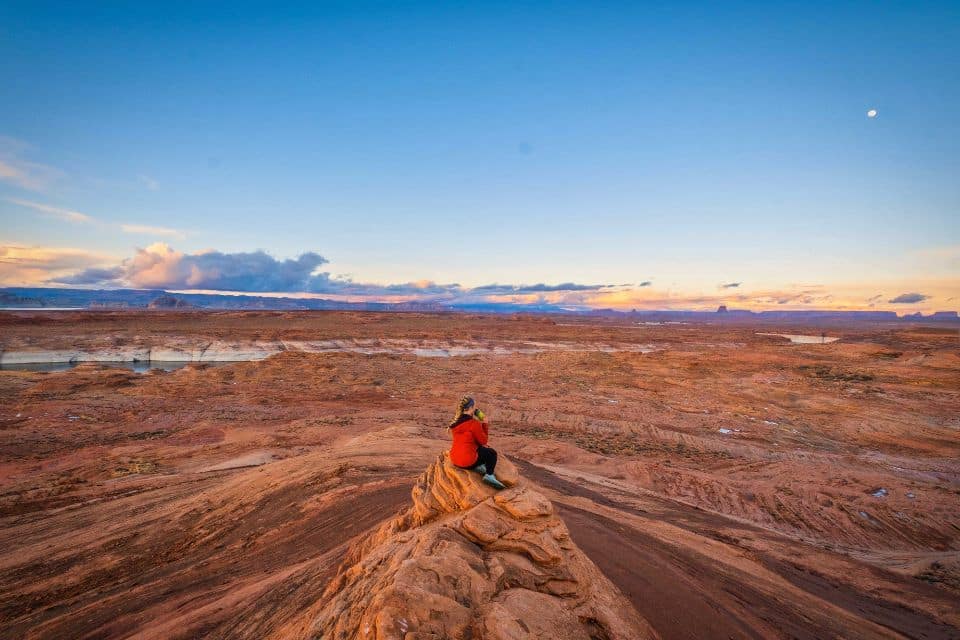 A person in a red jacket seated on a sandstone formation overlooking a vast desert landscape under a twilight sky, illustrating 'Hiking Tips for Beginners' on desert hiking essentials.