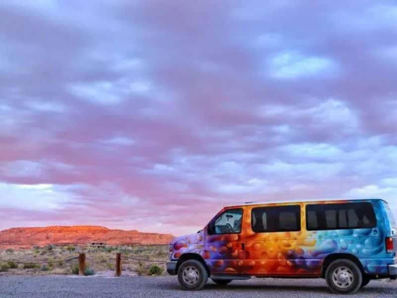 A colorful campervan parked in a scenic desert landscape with a dramatic sky at sunset, featuring hues of pink and purple.