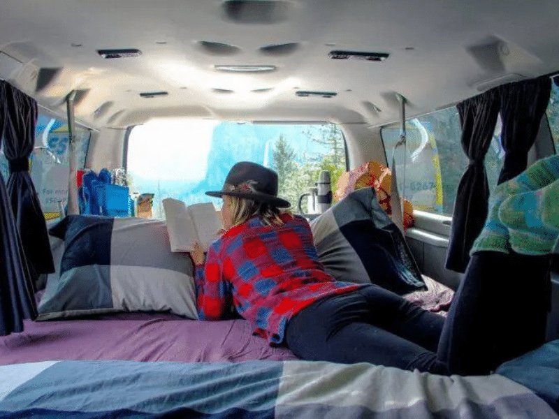 Interior view of a campervan with a person in a plaid shirt and hat lying on the bed, reading a book, with a picturesque view of a mountain and forest through the back windows.