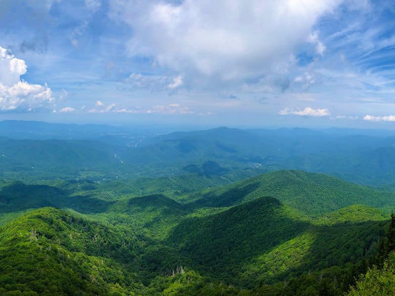 A panoramic view of lush green mountains under a bright blue sky with scattered clouds. The expansive landscape showcases the rolling hills and valleys covered in dense forest.
