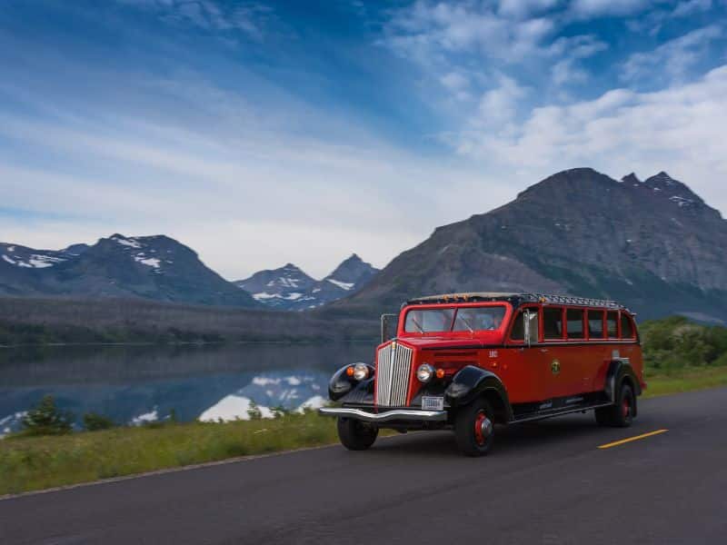 A vintage red tour bus driving along a scenic road with a lake and mountains in the background. The clear sky and majestic peaks reflect on the calm water, creating a picturesque backdrop.