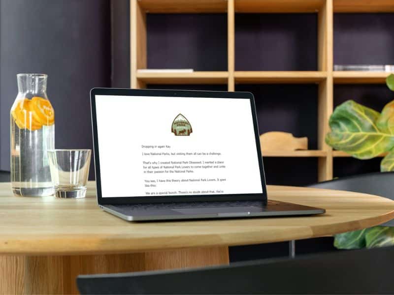 A laptop on a wooden table with an open page featuring National Park Obsessed logo and text about the love for national parks. Next to the laptop is a glass pitcher with orange slices and a glass of water.