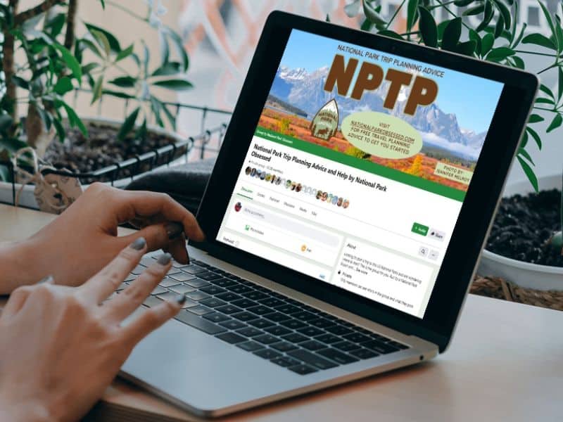 a laptop at a desk, with the screen displaying the Facebook group "National Park Trip Planning Advice and Help by National Park Obsessed" page. The surroundings include green plants and a window, creating a cozy, natural setting.