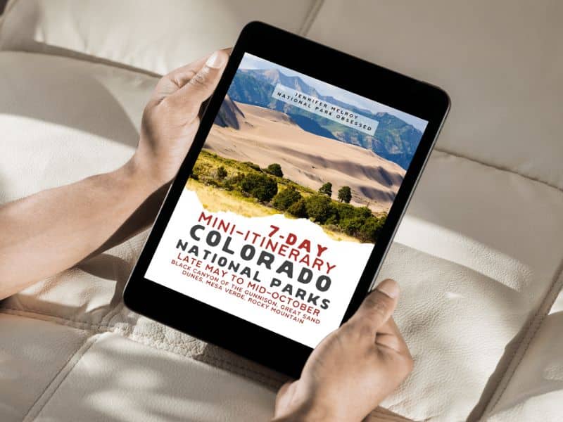 Hands holding a tablet displaying a 7-day mini-itinerary for Colorado National Parks, including an image of sand dunes and mountains in the background.